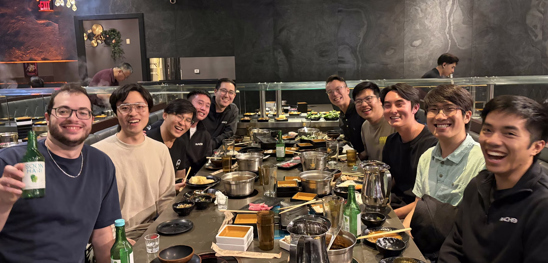 The team around a long hot pot table, one raising a bottle of soju